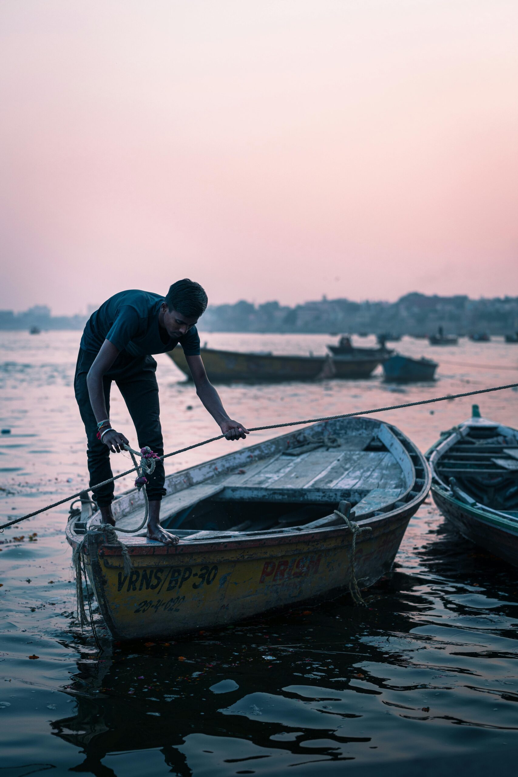 A man adjusts a boat on the Ganges River at Varanasi against a serene sunrise backdrop.